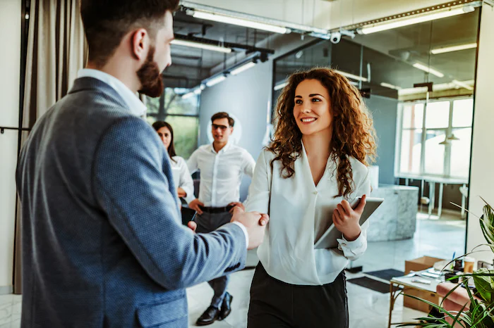 Businessman and businesswoman shaking hands in an office with colleagues in the background.