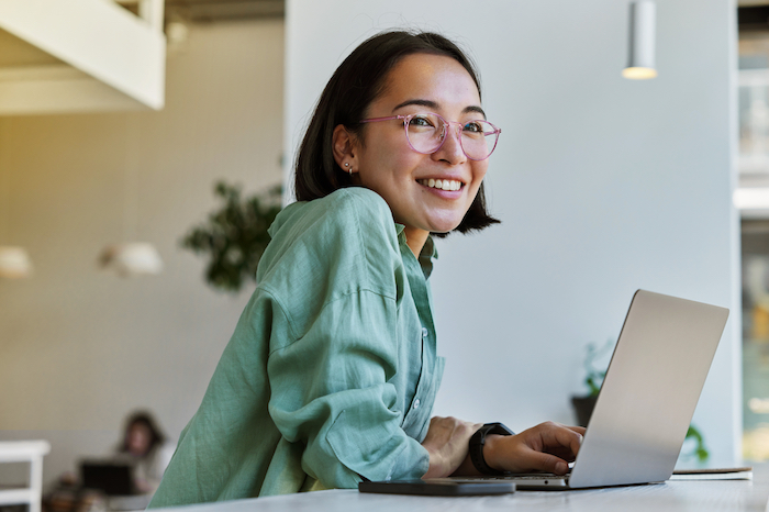 Woman with glasses and green shirt smiling while using a laptop in a café