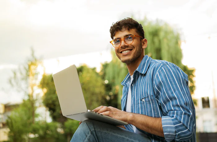 Smiling man in glasses using a laptop while sitting outside in a park