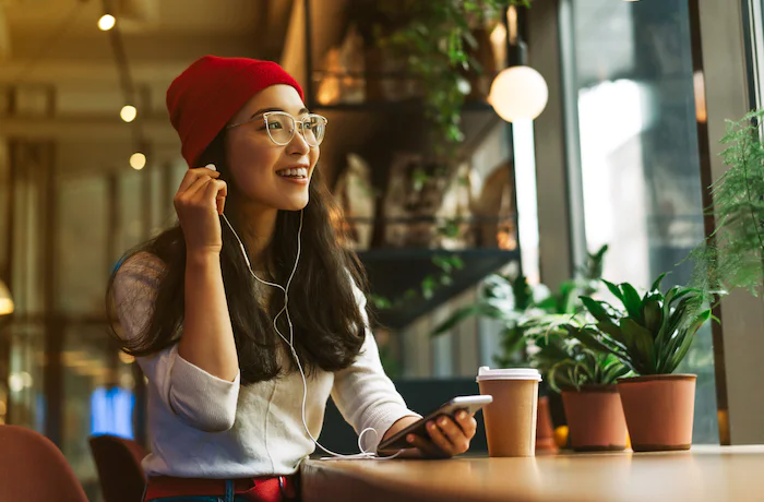 Woman wearing glasses and a red beanie listening to music and holding a smartphone at a café.