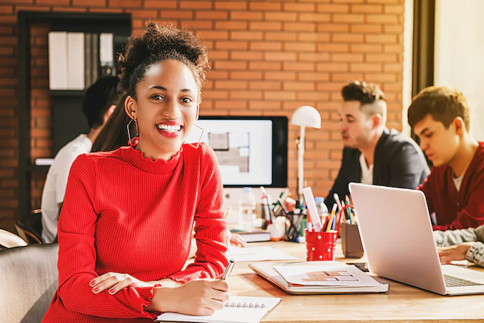 Woman in red sweater smiles and writes at a desk with colleagues working in the background.