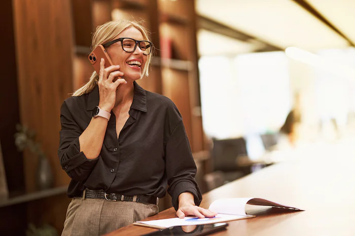 A woman with glasses smiles while talking on her phone in an office setting.