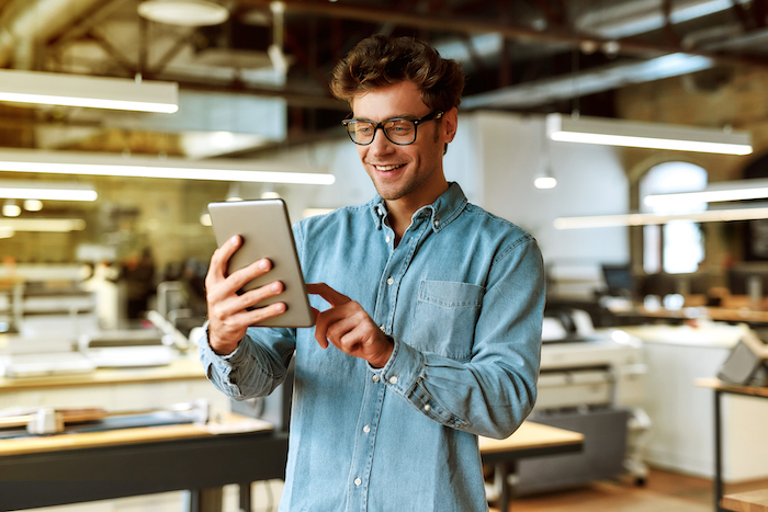 Man wearing glasses and denim shirt using a tablet in a contemporary office space.