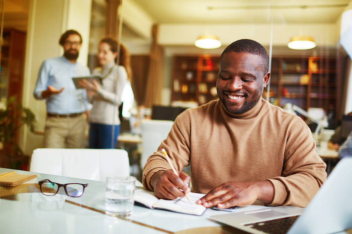 Man happily writes in a notebook at a desk in an office with two colleagues talking in the background.
