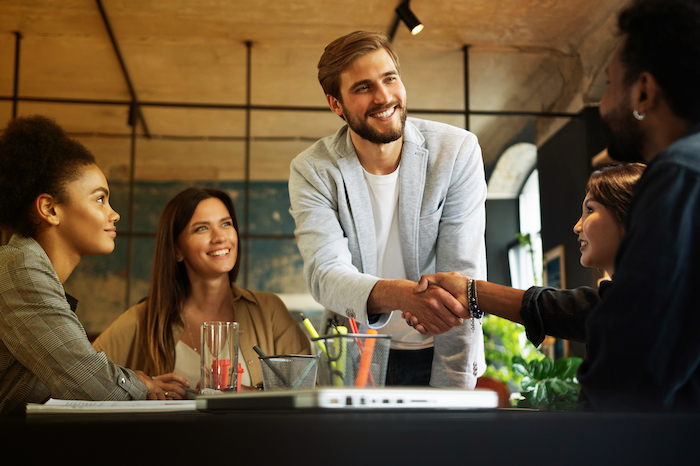 Group of diverse colleagues in a meeting, with two men shaking hands and three others smiling.