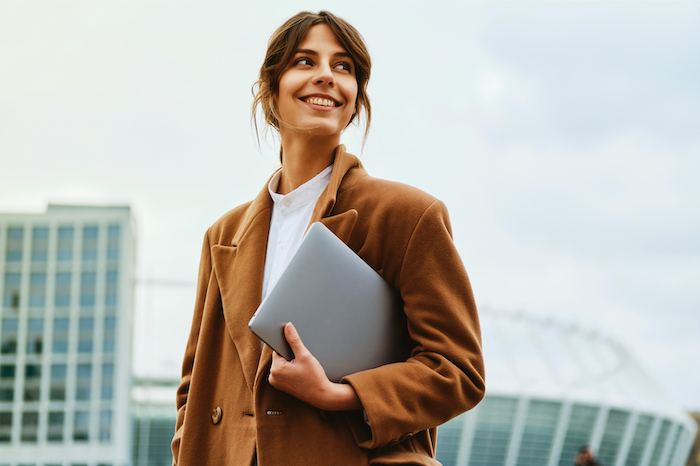 A young woman holds a laptop, smiles, and stands outdoors wearing a brown coat.