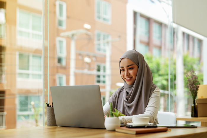 Woman in hijab working on a laptop at a desk near a large window in a modern office.