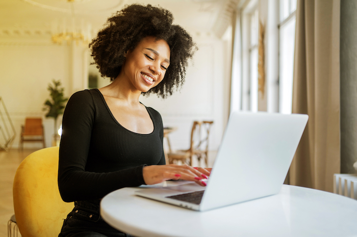 Smiling woman in black top typing on a laptop at a table in a bright, cozy café.