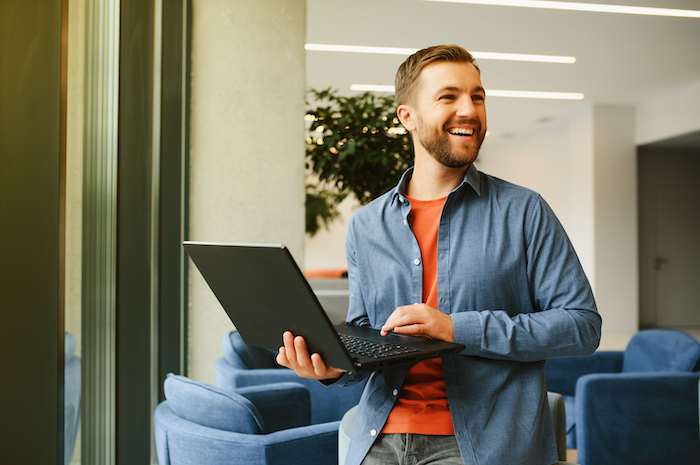 Man standing, smiling, using a laptop in a modern office space with blue chairs and large windows.