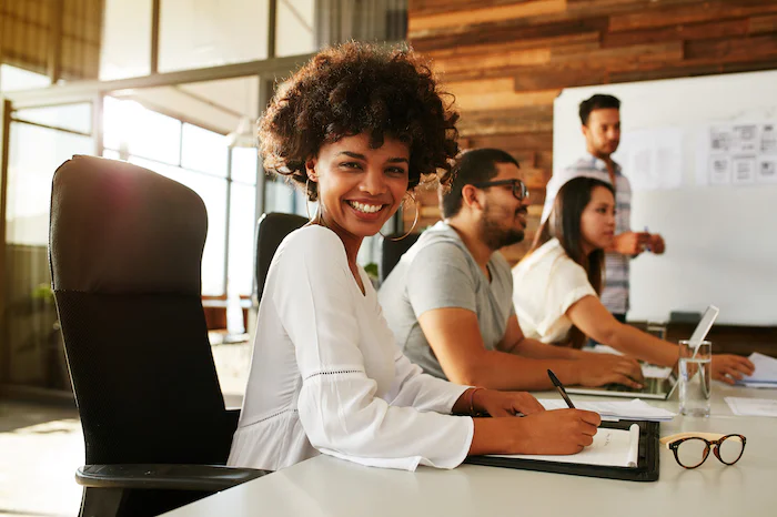 Smiling woman in white shirt sits at desk with colleagues working in modern office.