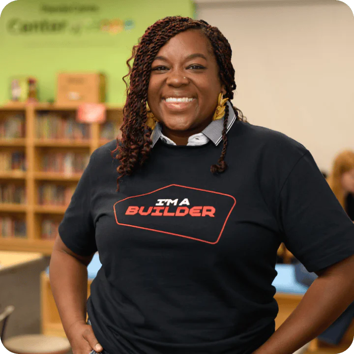 Woman with braided hair, wearing an I'm a Builder shirt, stands smiling in a library.