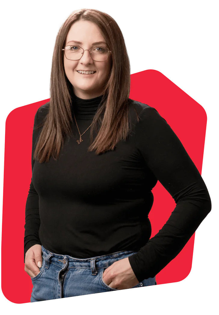 Woman with long brown hair, wearing glasses, a black turtleneck, and jeans, posing with hands in pockets.
