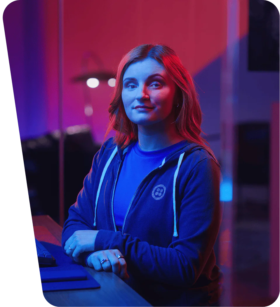 Woman in blue hoodie sits at a desk with neon lighting in background.