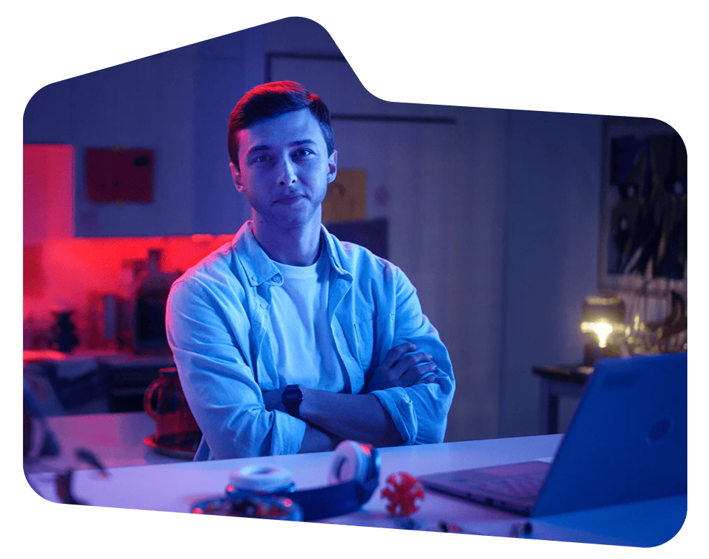 Man seated at desk with arms crossed in front of a laptop and various items in a modern, dimly lit room.