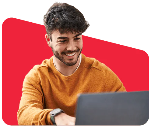 Young man in a mustard sweater smiling while using a laptop against a red background.