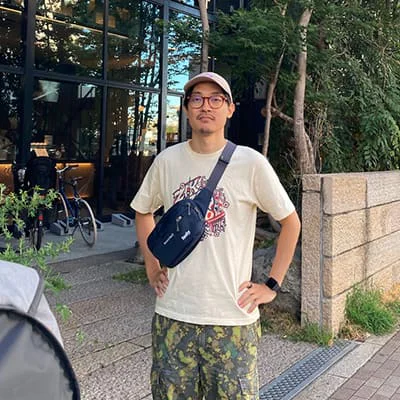 Man in glasses and a cap stands with his hands on his hips, wearing a beige shirt and camo shorts.