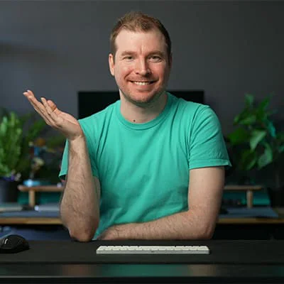 Man in a turquoise shirt smiling and gesturing with one hand at a desk with a keyboard and mouse