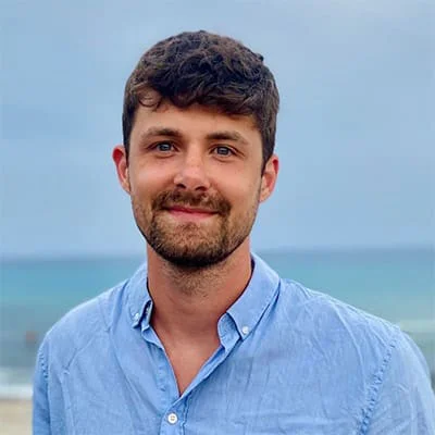 Man with short brown hair and beard wearing a blue shirt standing on a beach with the sea in the background