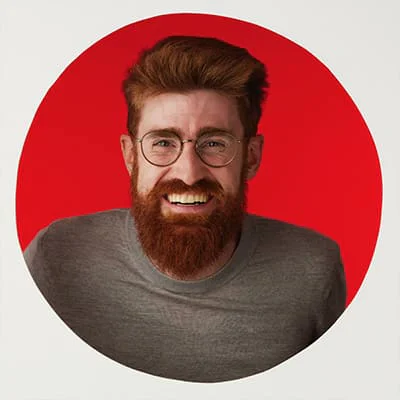 Man with red hair and beard, wearing glasses and a gray shirt, smiling in front of a red circle background.