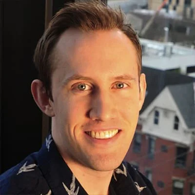 Man with short hair smiling, wearing a dark shirt with a bird pattern, with cityscape in background.