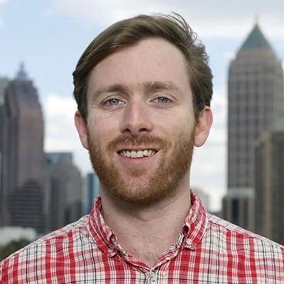 Man with beard in red plaid shirt smiling, standing in front of a city skyline with tall buildings.