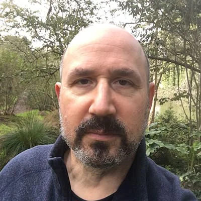 A man with a beard stands outside with trees and greenery in the background.