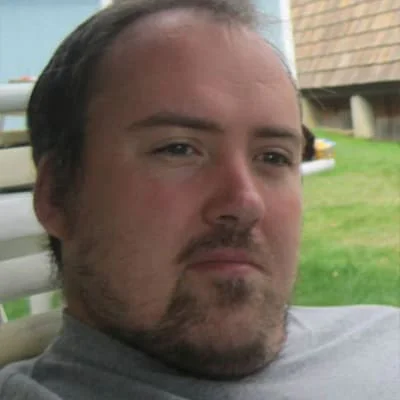 A man with a beard and mustache sitting on a bench outdoors with a slight smile on his face.