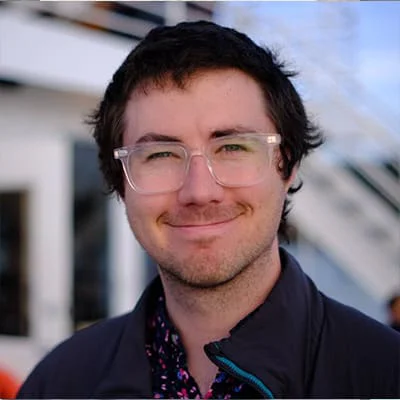 Person with black hair and clear glasses smiling in an outdoor setting with a building in the background.