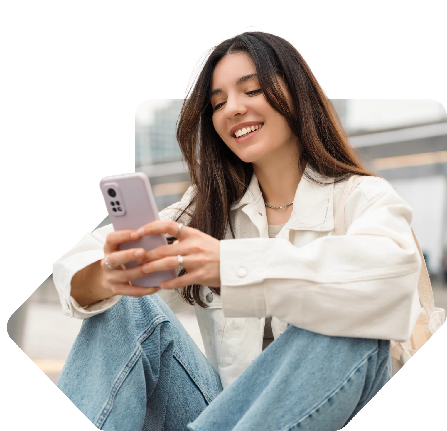 Woman sitting outside, smiling and using her smartphone, wearing a white jacket and blue jeans.