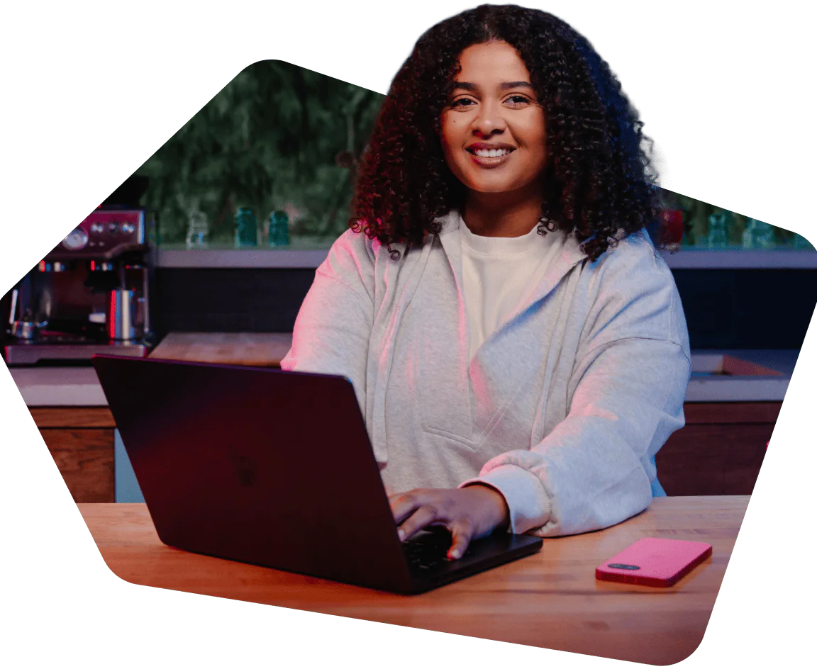 Woman with curly hair using a laptop at a wooden table, coffee machine in the background.