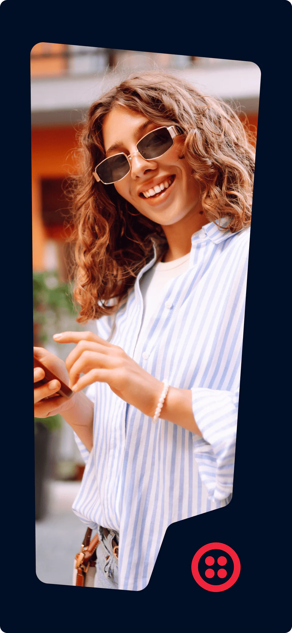 Woman with curly hair and sunglasses smiling while using her smartphone outdoors.