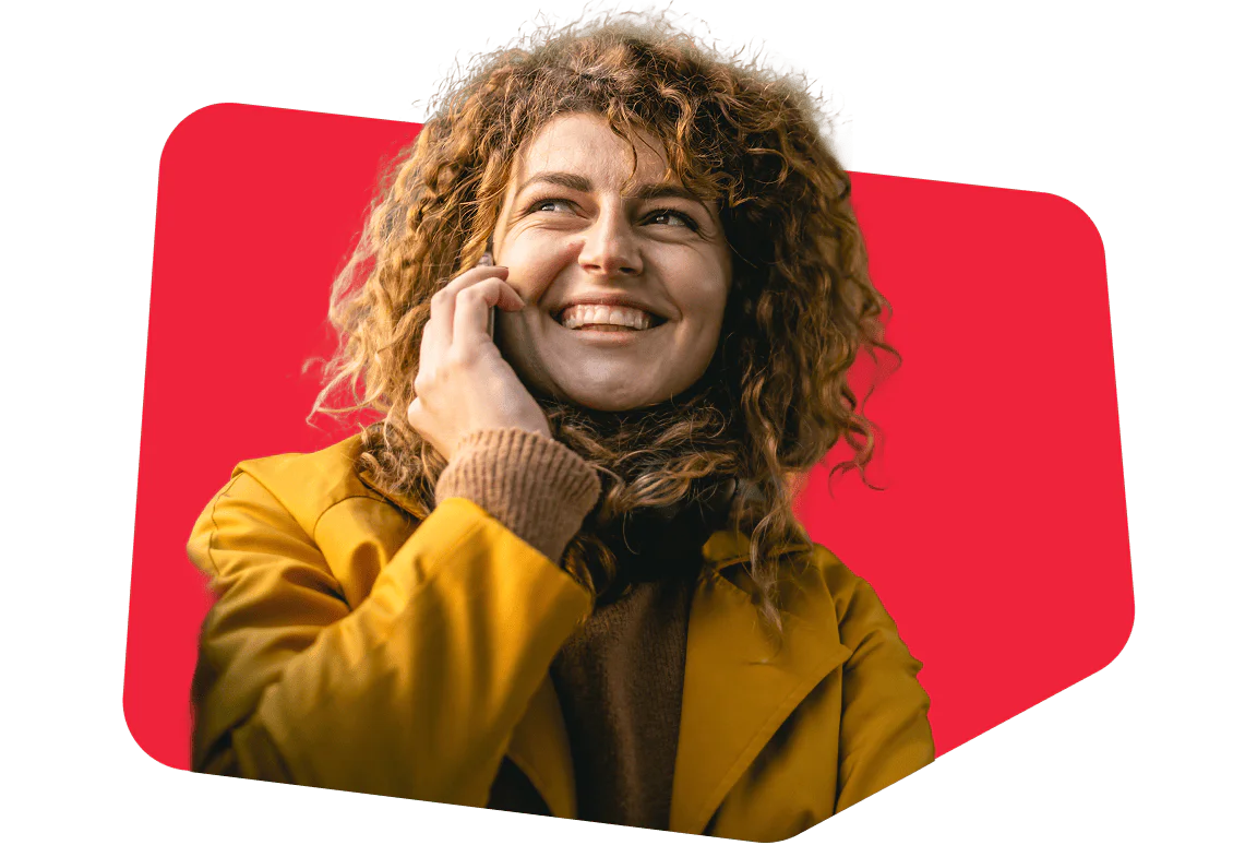 Woman with curly hair and yellow coat talking on phone, smiling against a red background.