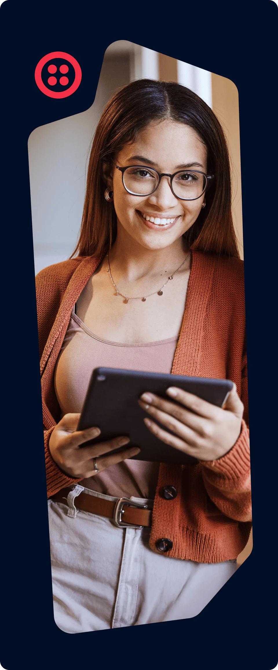 A smiling woman with glasses and long hair holds a tablet, wearing a brown cardigan and beige pants.