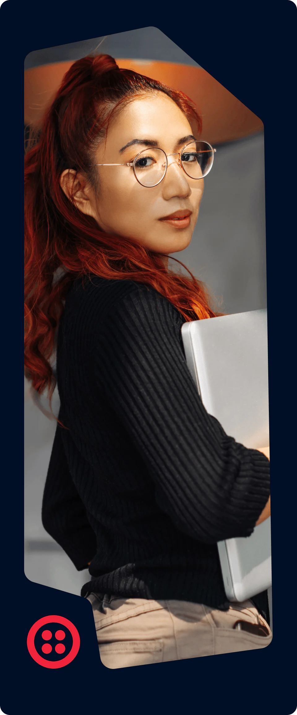 Woman with red hair and glasses holding a laptop, looking confidently at the camera.