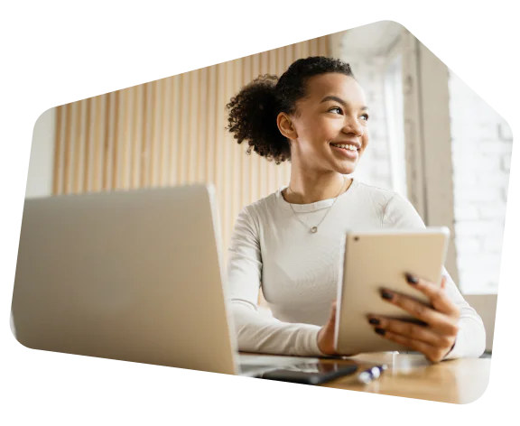 Smiling woman holding a tablet and sitting in front of a laptop in a bright, modern workspace.