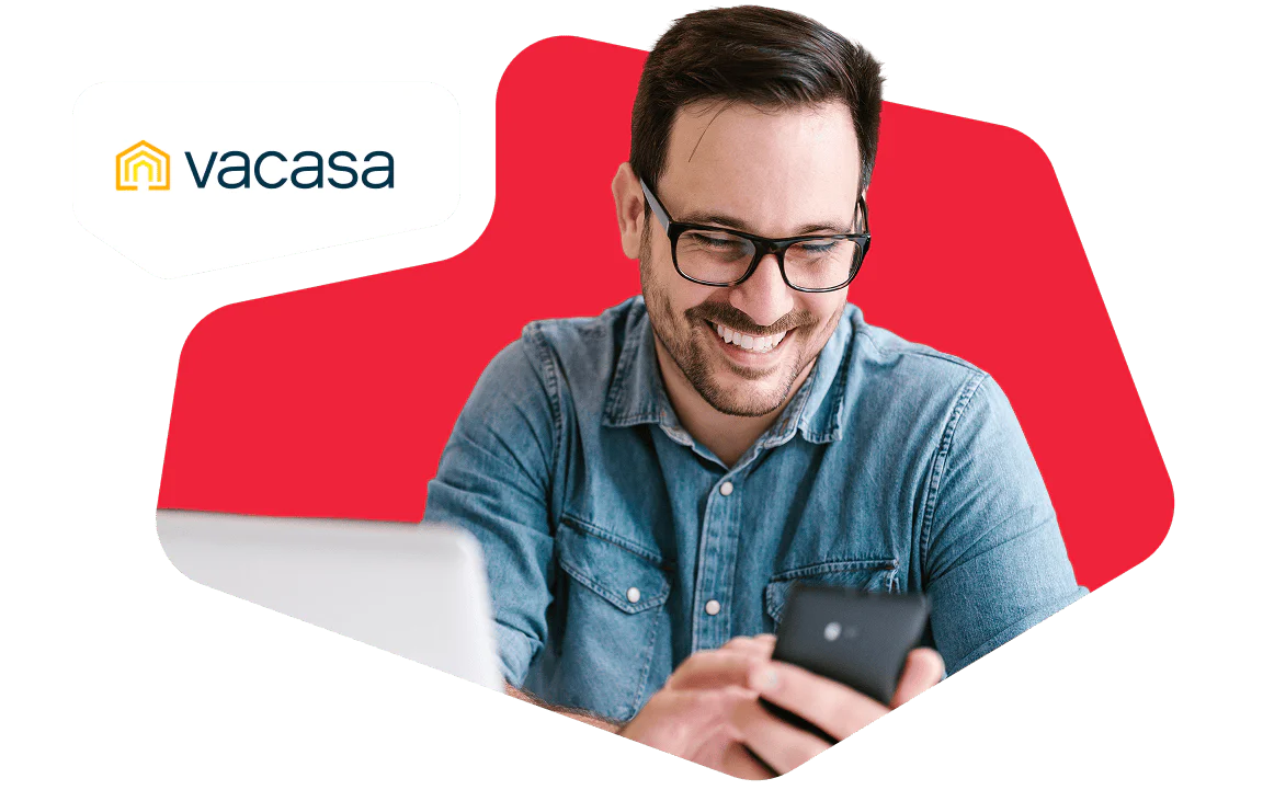 Smiling Man Using Smartphone and Laptop at Work Man smiling while looking at his smartphone, with a laptop in front of him and a Vacasa logo.