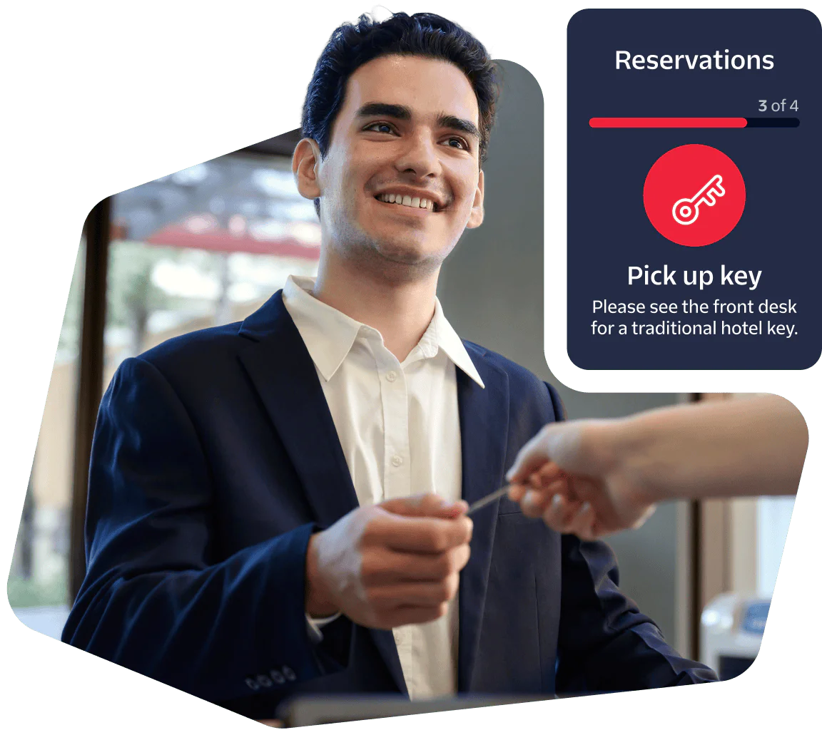Smiling hotel receptionist gives a room key to a guest with reservation confirmation details on-screen.