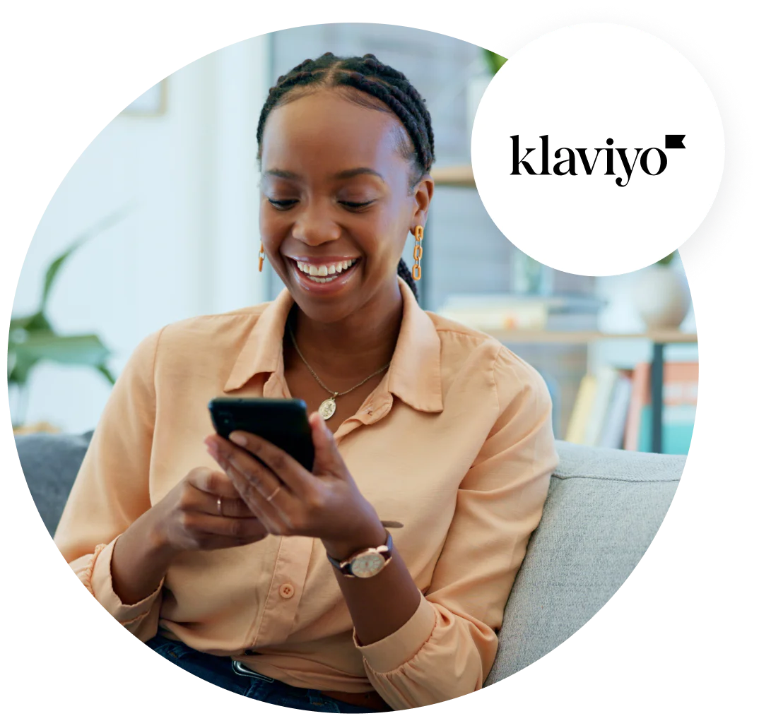 Woman smiling while looking at her smartphone, seated on a couch with a Klaviyo logo in the top right corner.