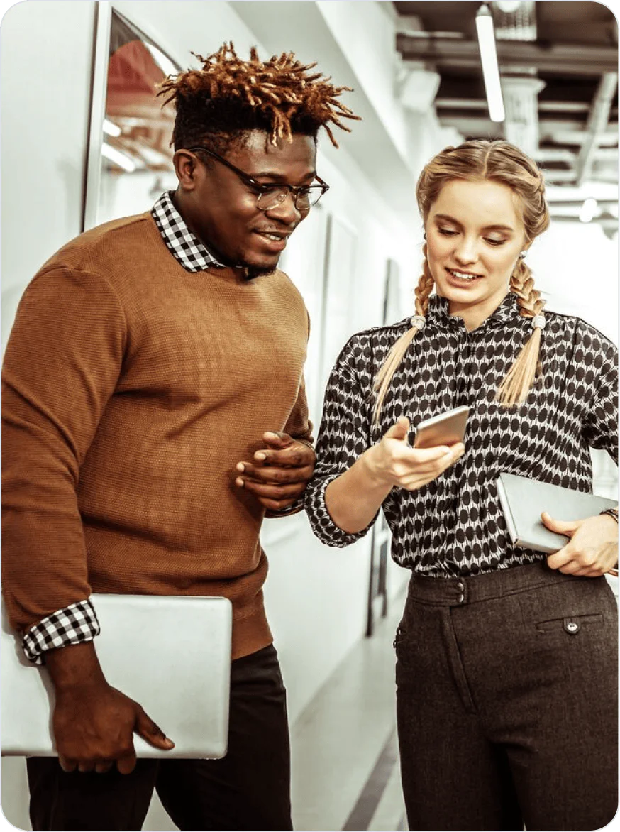 Two colleagues looking at a smartphone and discussing something while standing in a modern office corridor.