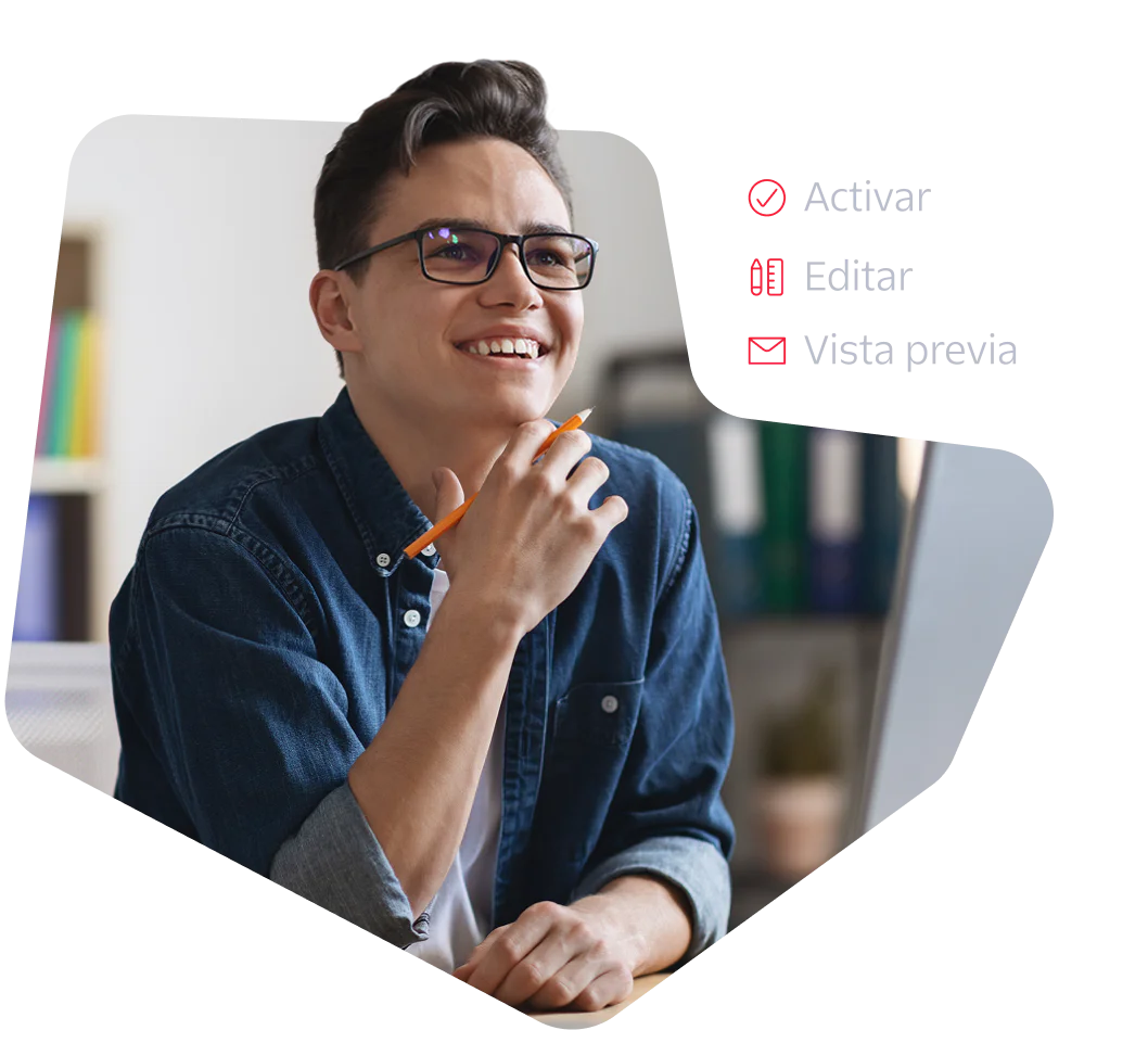 A young man with glasses and a denim shirt smiles while working at a computer.