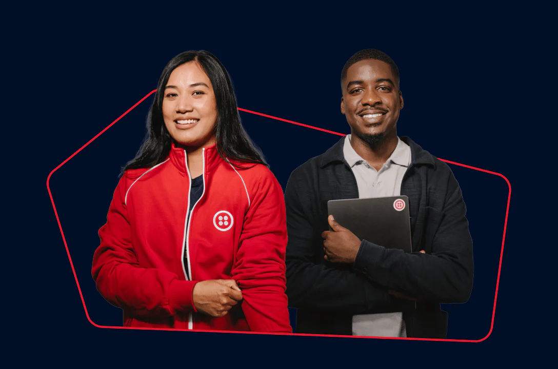 Two smiling individuals, one holding a laptop, wearing branded clothing against a dark background.